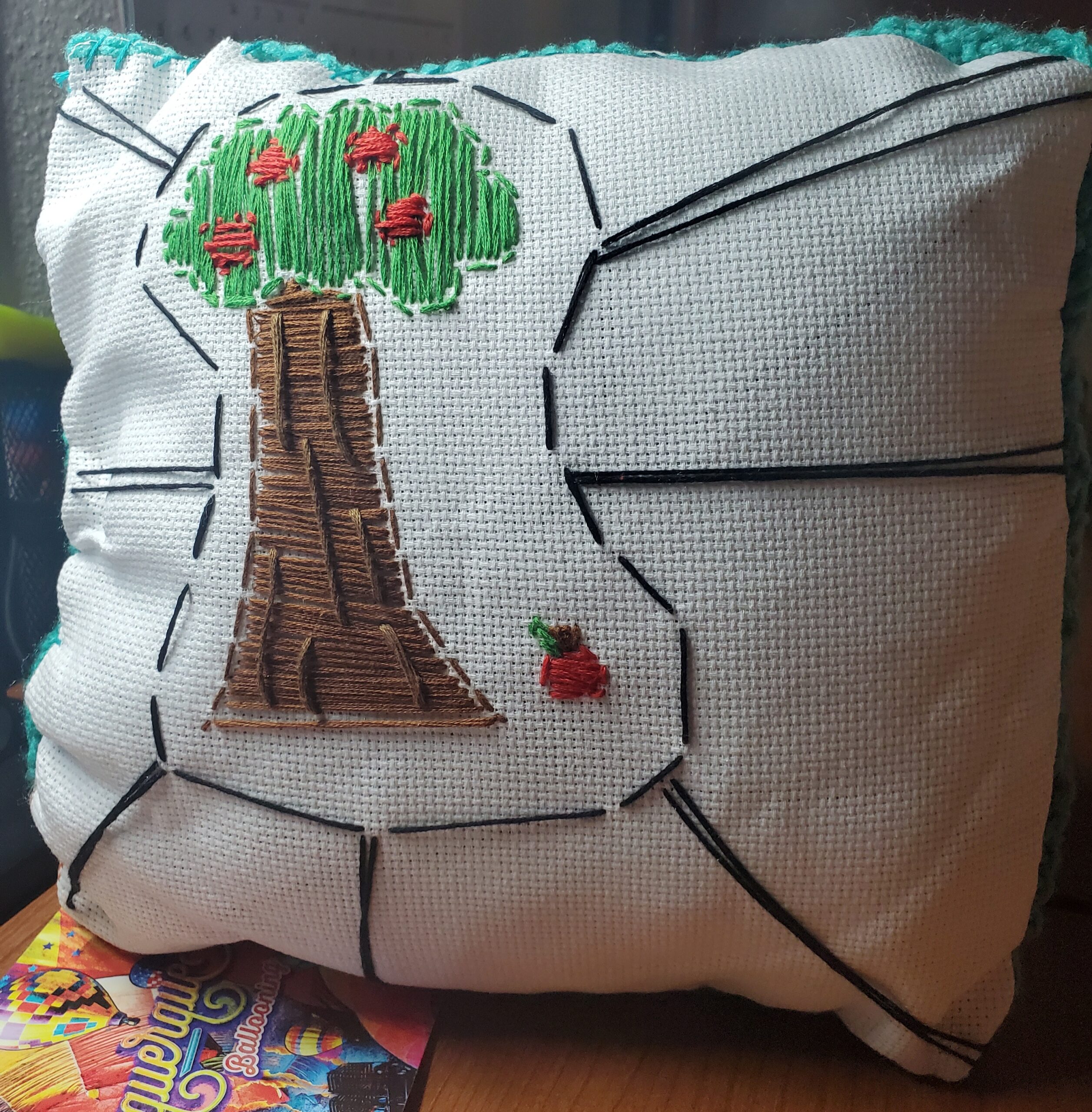 a white pillow with an embroidered apple tree on it. One apple is on the ground beside the tree.