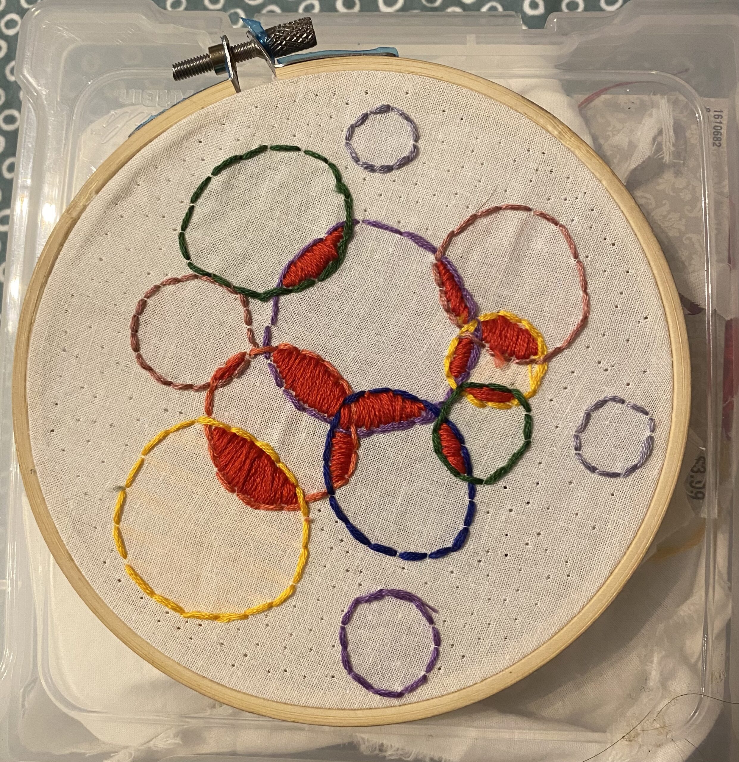 White fabric, dotted with tiny, deliberate holes poked through it is held in an embroidery hoop. In red, blue, purple, yellow, and green embroidery floss are several circles backstitched in different colors. Where the circles meet and overlap, the area is filled with red embroidery floss in a satin stitch pattern.